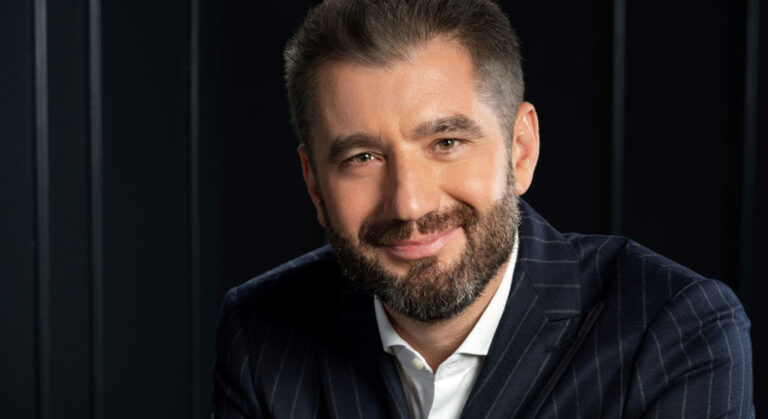 Portrait of a smiling man with a beard in a navy pinstripe suit and white shirt against a dark background, a professional headshot.